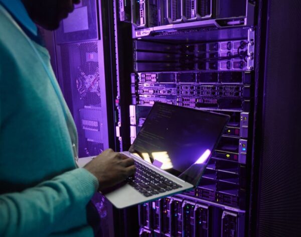 cropped-shot-african-american-data-engineer-holding-laptop-while-working-with-supercomputer-server-room-lit-by-blue-light-copy-space-min