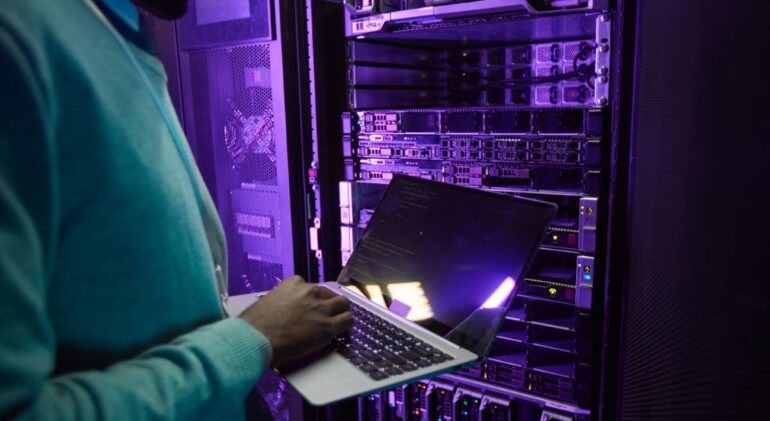 cropped-shot-african-american-data-engineer-holding-laptop-while-working-with-supercomputer-server-room-lit-by-blue-light-copy-space-min